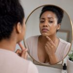 mulher negra olhando-se no espelho tocando o rosto com expressão frustrada pensando em como diminuir a oleosidade da pele